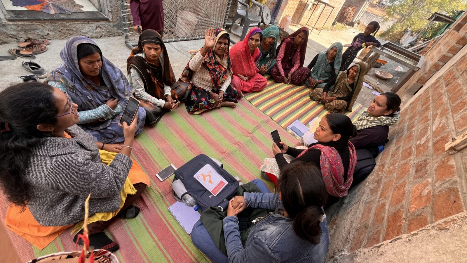 A women’s self-help group meeting. The moderator holds a phone; some members raise a hand, others hold phones.